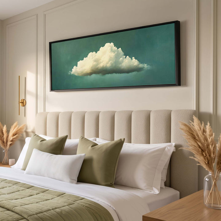 A framed wall art print depicting a fluffy white cloud against a teal-green sky, displayed above a beige upholstered headboard in a styled bedroom setting.
