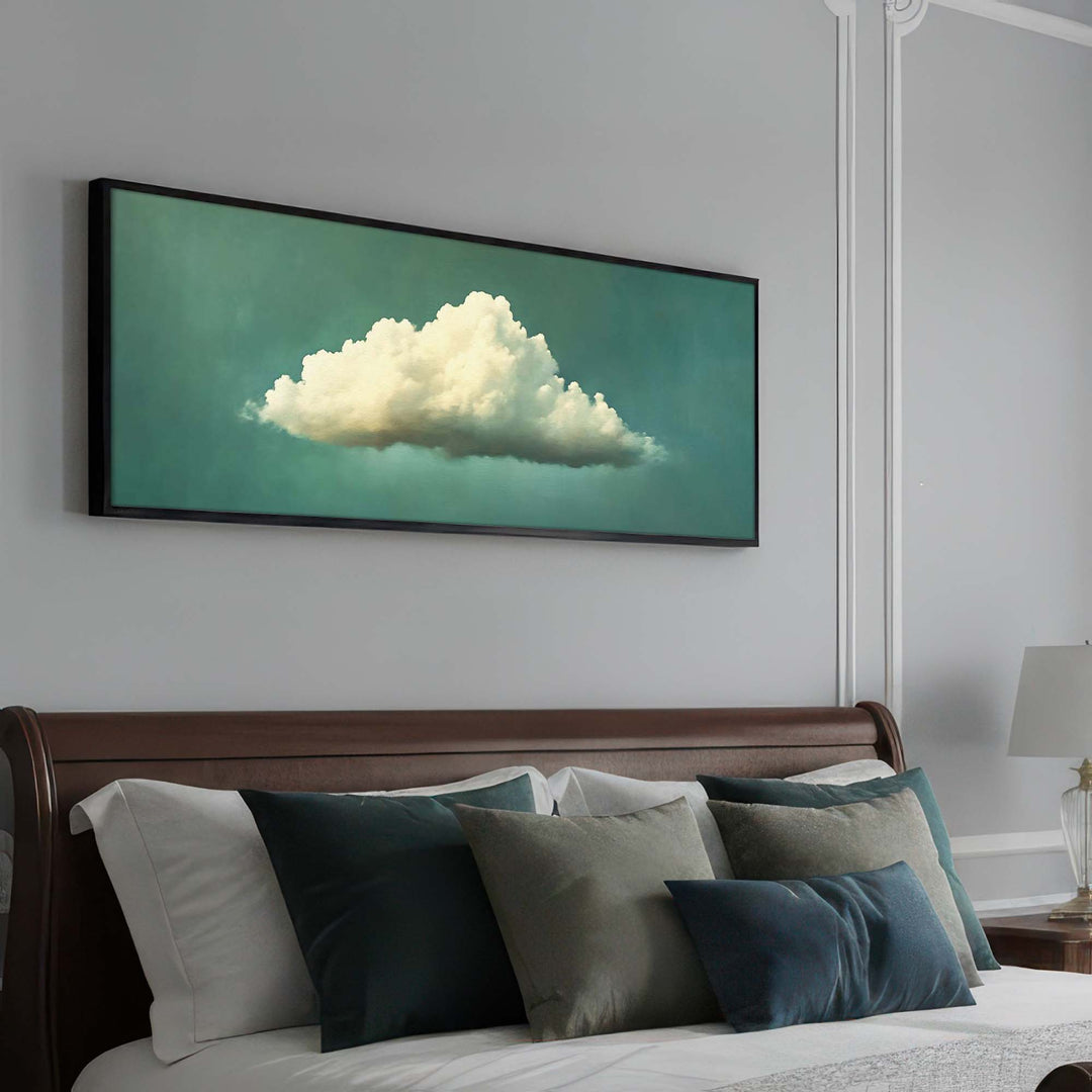 A horizontal framed wall art print featuring a large white cloud against a teal-green background, displayed above a bed with white and gray pillows in a bedroom setting.
