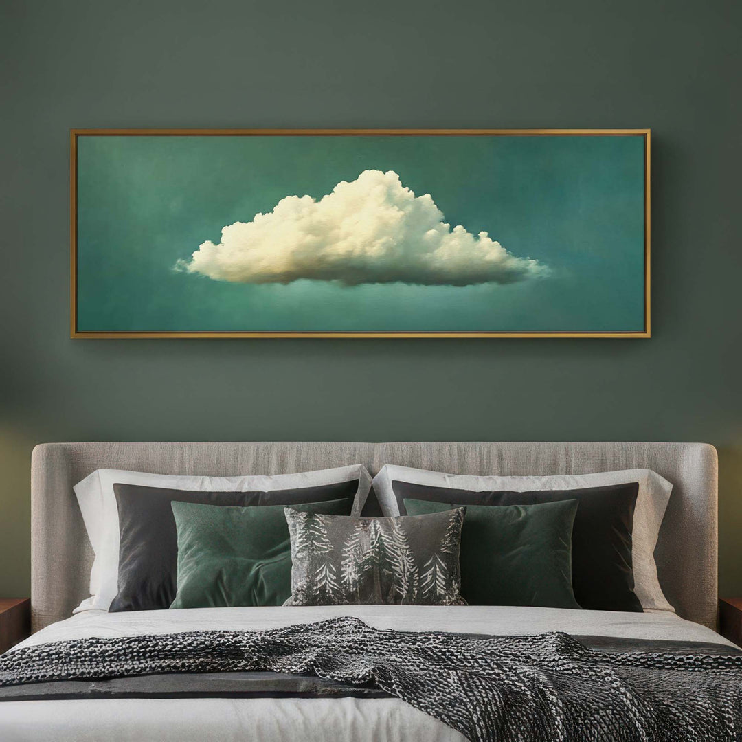 A framed wall art print depicting a large cream-colored cloud against a teal-green background, hung above a neatly made bed with dark green and gray pillows in a modern bedroom setting.