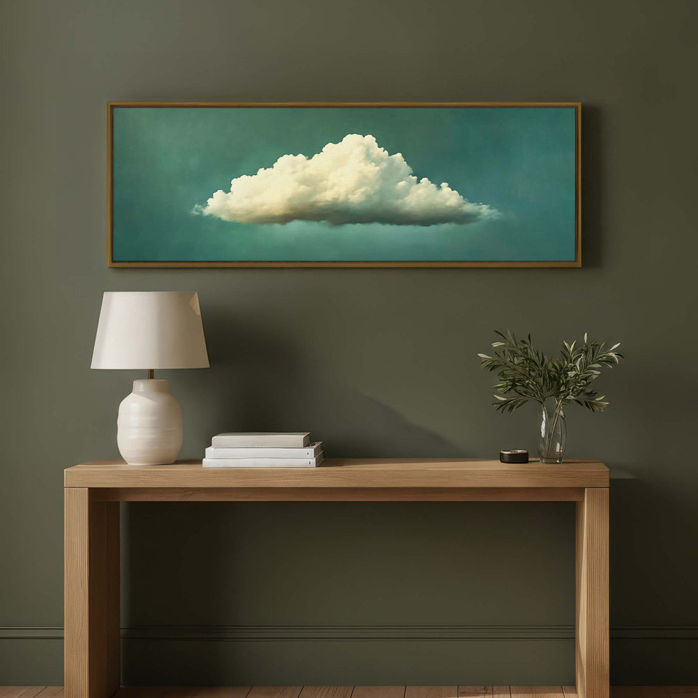 A framed horizontal canvas print of a white cloud against a teal background, displayed on a dark olive green wall above a light wood console table with a white lamp and a small plant.