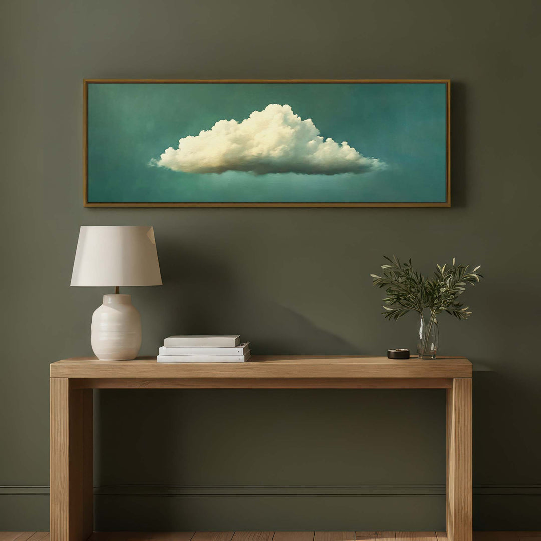 A framed horizontal canvas print of a white cloud against a teal background, displayed on a dark olive green wall above a light wood console table with a white lamp and a small plant.