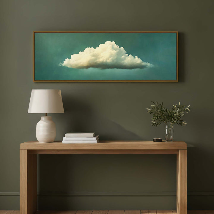 A framed horizontal canvas print of a white cloud against a teal background, displayed on a dark olive green wall above a light wood console table with a white lamp and a small plant.