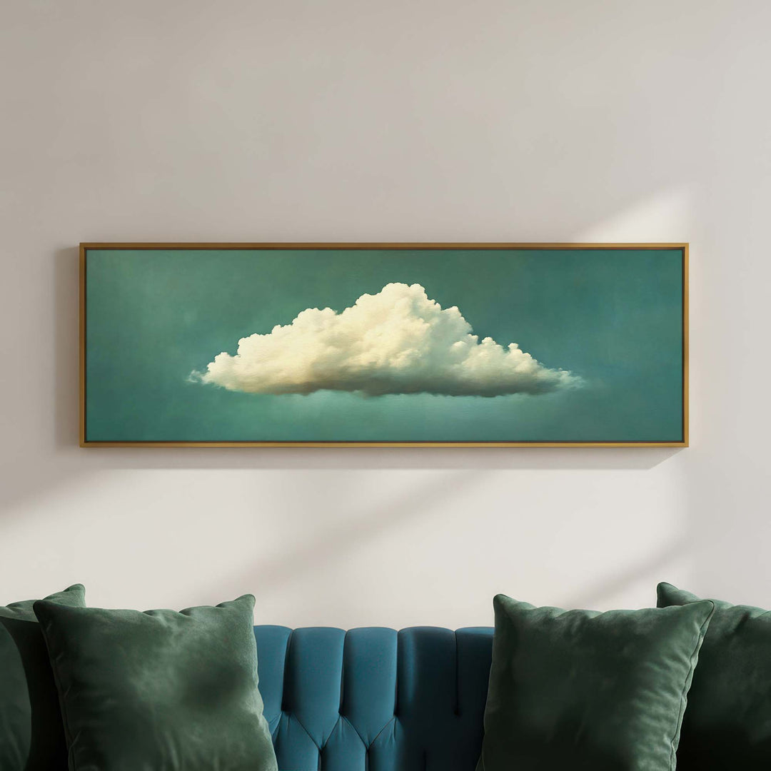 A framed horizontal canvas print of a fluffy white cloud against a teal sky, displayed on a light gray wall above a teal tufted sofa with green pillows.