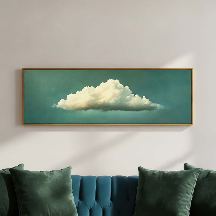 A framed horizontal canvas print of a fluffy white cloud against a teal sky, displayed on a light gray wall above a teal tufted sofa with green pillows.
